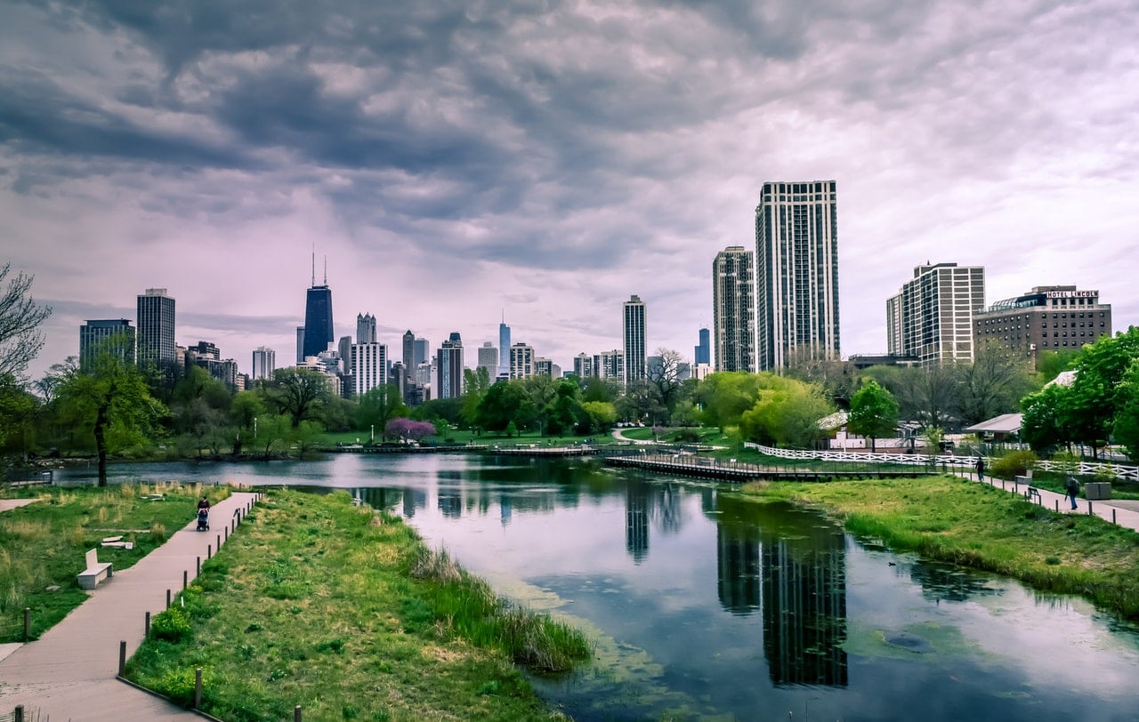  An aerial view of a bustling city with green spaces, including a park with a pond and a skyline with many skyscrapers.