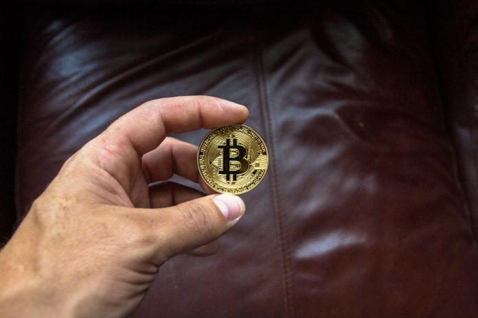 hand holding gold coin with bitcoin logo above a brown leather couch
