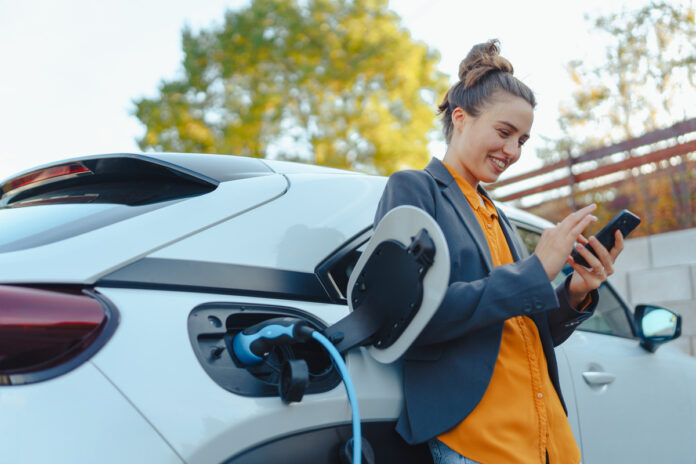 A smiling young woman in a blazer stands outside using her cell phone and charging her electric vehicle.