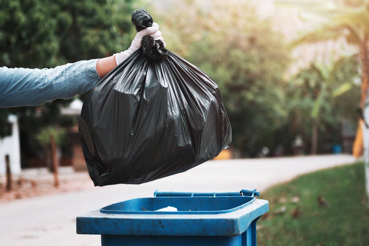 A person wearing a white latex glove throws a tied black plastic garbage bag into a blue trash bin outdoors.