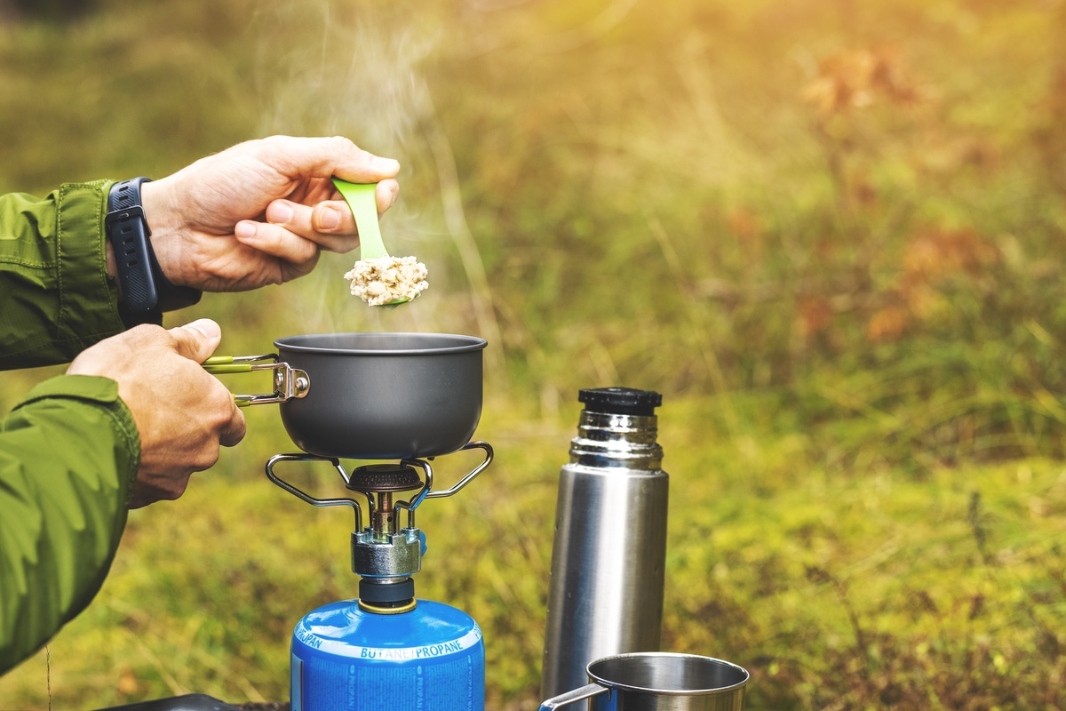 Outside, a person in a green rain jacket holds a green spoon of steaming oats over a pot on a gas burner.